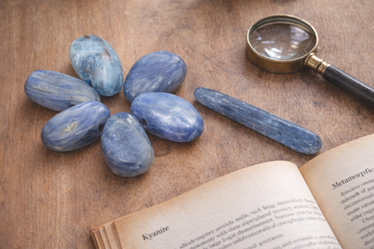 Blaue Kyanit-Trommelsteine und Rohkristall mit seidiger Streifenstruktur auf warmem Holztisch mit Lupe und Mineralogie-Buch